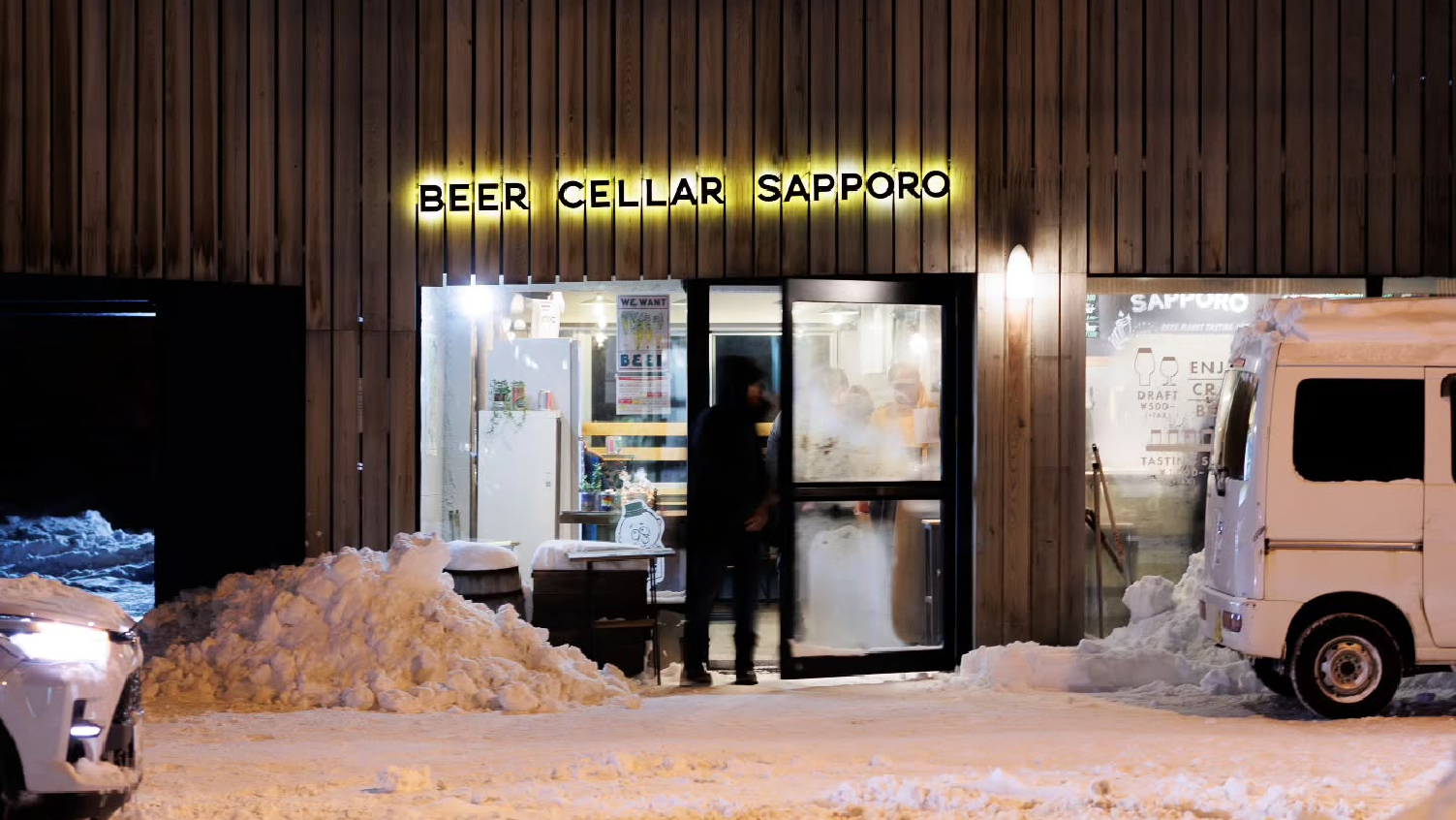 Photo: Beer Cellar Sapporo storefront. Snow is piled high around the sides of the entrance and two people are entering through the foggy entrance doors.
