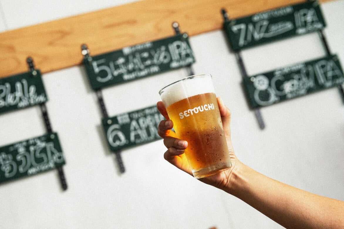 A hand holds a full pint glass of beer that has the wordmark Setouchi printed on it. In the background is a wall menu composed of black numbered signs with different beers listed on them.