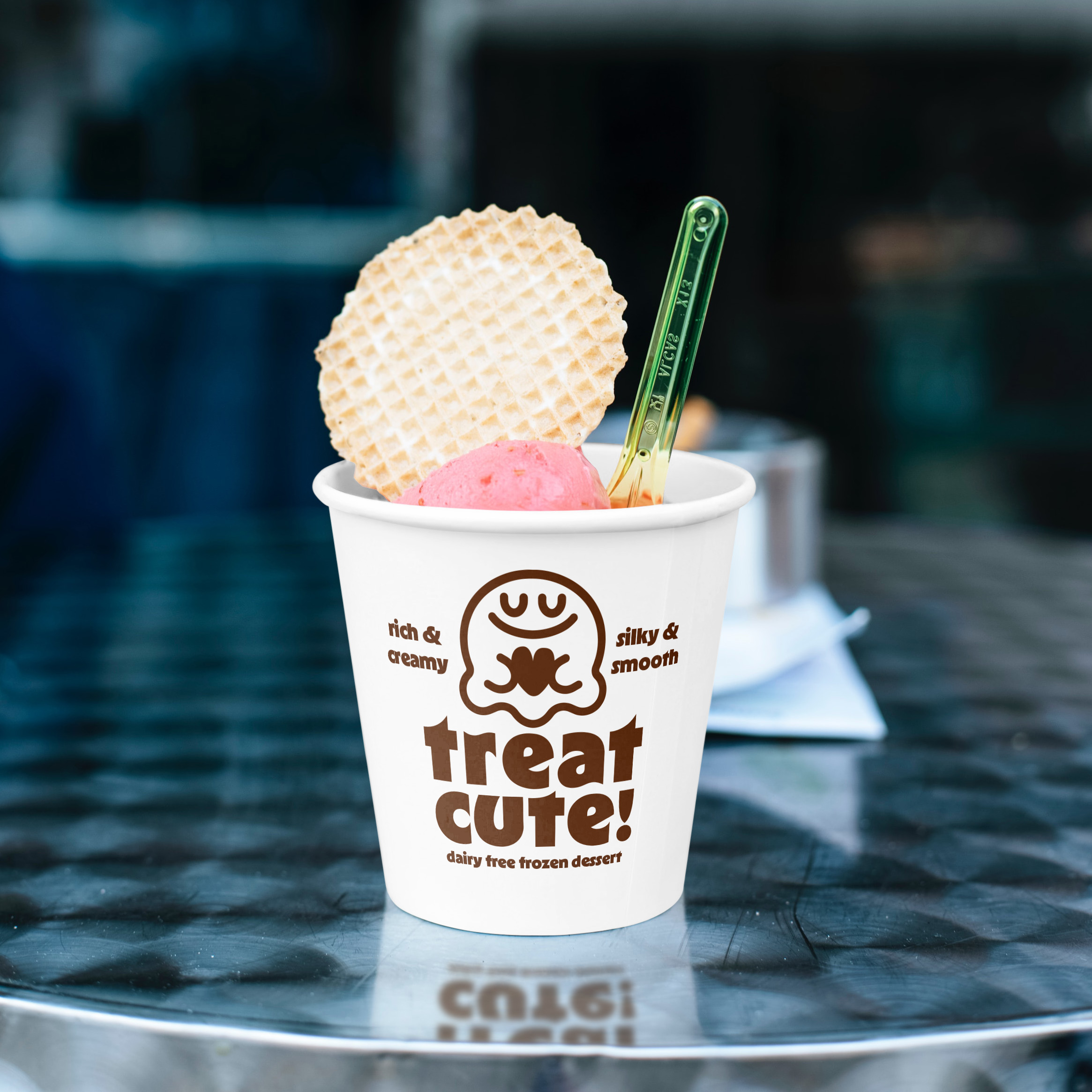 A white cup with a scoop of pink ice cream inside, sitting on a reflectiv metal cafe table. A thin waffle cookie and a clear spoon are sticking out of it.